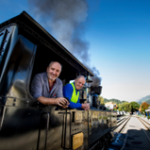 Zwei Männer lehnen sich aus dem Fenster einer schwarzen Dampflokomotive, einer im gestreiften Hemd, der andere in einer blauen Uniform, im Hintergrund sind Gleise und Gebäude zu sehen.