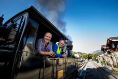 Zwei Männer lehnen sich aus dem Fenster einer schwarzen Dampflokomotive, einer im gestreiften Hemd, der andere in einer blauen Uniform, im Hintergrund sind Gleise und Gebäude zu sehen.