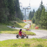 An einem sonnigen Tag fahren Menschen mit dreirädrigen Go-Karts einen gewundenen Schotterweg durch ein Waldgebiet hinunter, während über ihnen ein Skilift zu sehen ist.