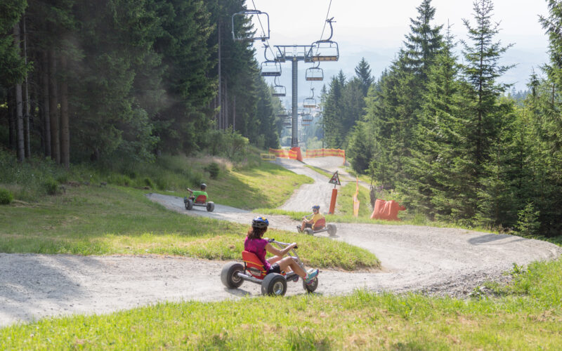 An einem sonnigen Tag fahren Menschen mit dreirädrigen Go-Karts einen gewundenen Schotterweg durch ein Waldgebiet hinunter, während über ihnen ein Skilift zu sehen ist.