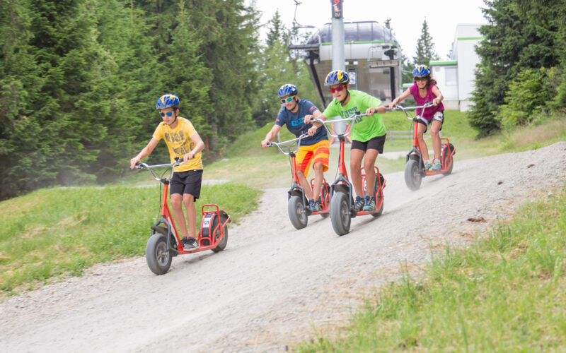 Vier Personen mit Helmen fahren mit großen Rollern einen Kiesweg in einem bewaldeten Außenbereich in der Nähe eines Skilifts hinunter.