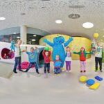 Kinder und Erwachsene stehen in einem Spielzimmer mit bunten Blöcken, einige halten Bälle, und in der Mitte posiert eine Person in einem blauen Monsterkostüm.