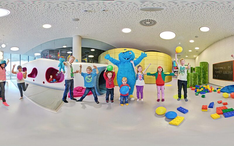 Kinder und Erwachsene stehen in einem Spielzimmer mit bunten Blöcken, einige halten Bälle, und in der Mitte posiert eine Person in einem blauen Monsterkostüm.