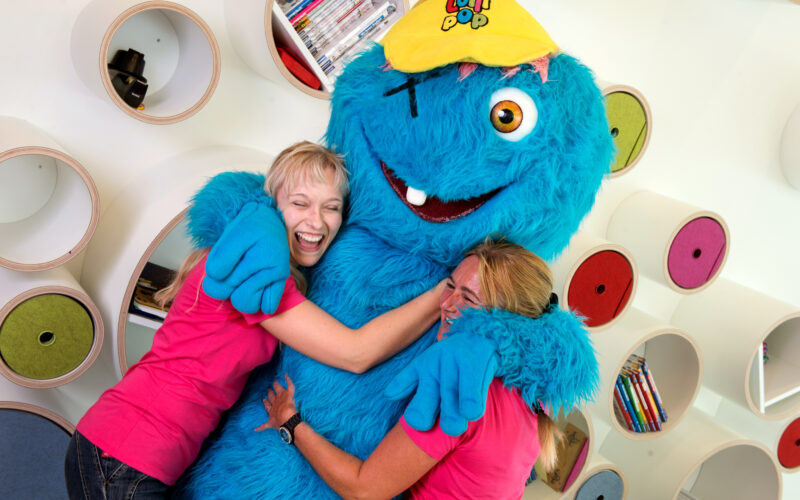 Zwei Frauen in rosa Hemden umarmen eine Person in einem großen blauen Monsterkostüm mit gelber Mütze, die vor runden Regalen mit Büchern und Spielen steht.