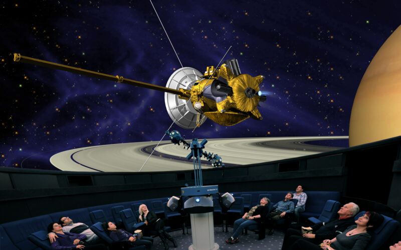 Die Besucher eines Planetariums sehen auf einem großen Kuppelbildschirm die Projektion eines Raumschiffs in der Nähe des Saturns und seiner Ringe.