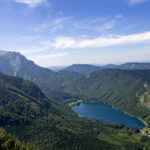 Luftaufnahme einer grün bewaldeten Berglandschaft mit einem blauen See im Tal unter einem klaren, teilweise bewölkten Himmel.