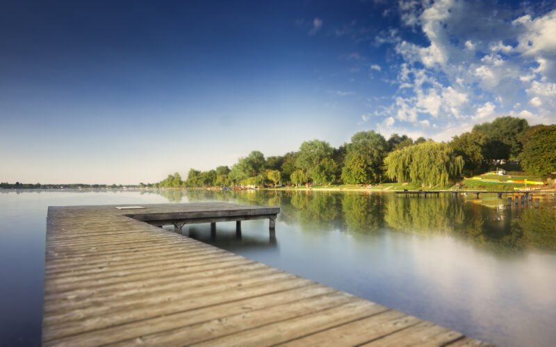 Der hölzerne Steg erstreckt sich über einen ruhigen See mit Bäumen und grasbewachsenen Ufern, die sich im Wasser spiegeln, unter einem blauen Himmel mit vereinzelten Wolken.