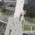 Menschen verschiedenen Alters klettern an einer großen künstlichen Kletterwand, die aus einem Schwimmbecken hervorgeht, während im Hintergrund Wasserrutschen und Bäume zu sehen sind.