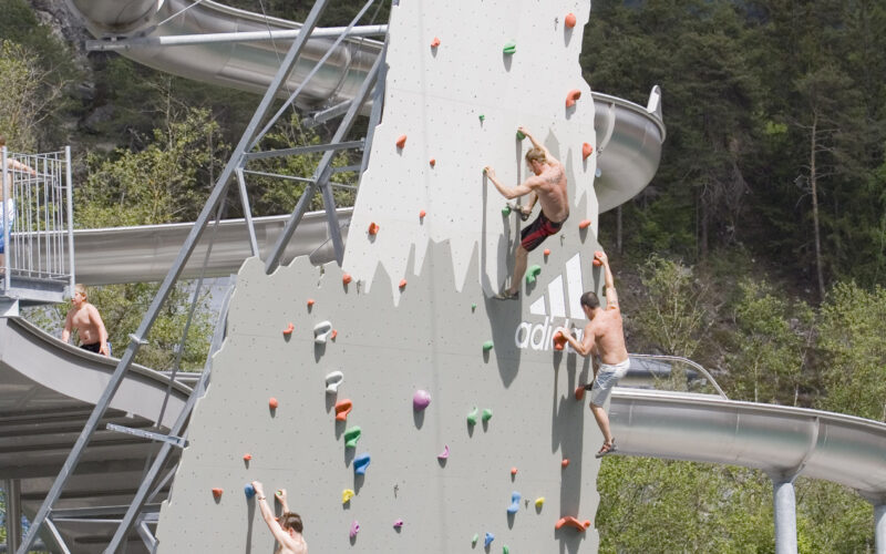 Menschen verschiedenen Alters klettern an einer großen künstlichen Kletterwand, die aus einem Schwimmbecken hervorgeht, während im Hintergrund Wasserrutschen und Bäume zu sehen sind.