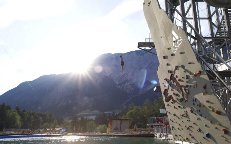 Eine Person springt von einer Kletterwand im Freien in einen See, mit Bergen und Bäumen im Hintergrund und der Sonne, die teilweise hinter den Gipfeln sichtbar ist.