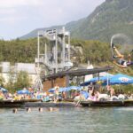 Eine Person schwingt sich an einer Seilrutsche in einen See, während die Leute zusehen und schwimmen; im Hintergrund sind eine Plattform am Wasser und Berge zu sehen.
