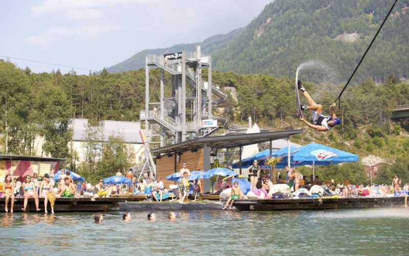 Eine Person schwingt sich an einer Seilrutsche in einen See, während die Leute zusehen und schwimmen; im Hintergrund sind eine Plattform am Wasser und Berge zu sehen.