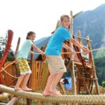 Zwei Kinder balancieren auf einer Seilbrücke auf einem Spielplatz im Freien mit Holzstrukturen, umgeben von Bäumen und Bergen.