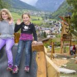 Zwei junge Mädchen sitzen auf einer Spielplatzrutsche im Freien, lächeln und winken in die Kamera, während im Hintergrund ein Dorf und Berge zu sehen sind.