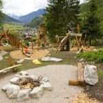 Spielplatz im Freien mit Holzkonstruktionen, Bänken und einer Feuerstelle, umgeben von Bäumen und mit Blick auf eine Berglandschaft und ein Dorf.
