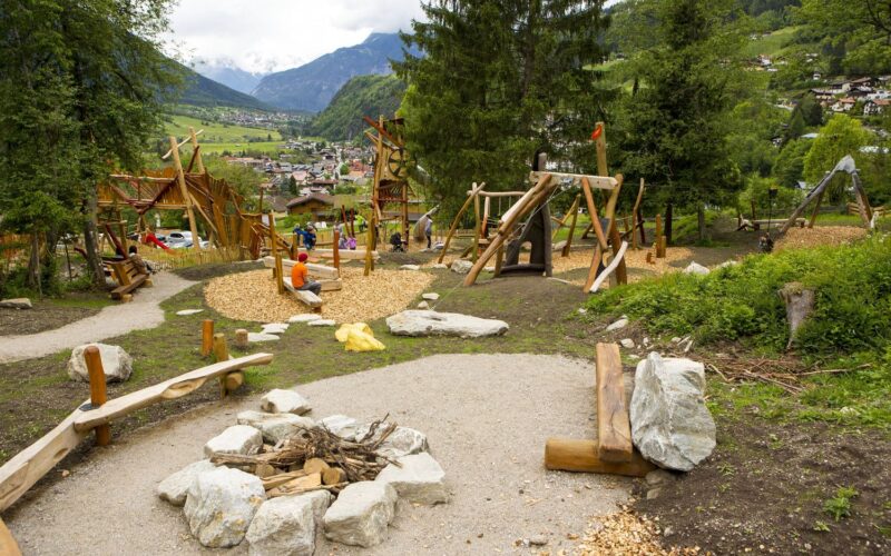 Spielplatz im Freien mit Holzkonstruktionen, Bänken und einer Feuerstelle, umgeben von Bäumen und mit Blick auf eine Berglandschaft und ein Dorf.
