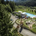 Luftaufnahme eines Freizeitgeländes mit Schwimmbad, Wasserrutsche, Spielplatz, Parkplatz und Bäumen in einer bergigen Landschaft.
