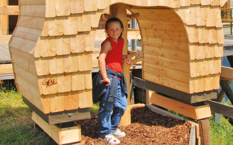 Ein Kind in rotem Hemd und blauer Hose steht an einem sonnigen Tag am Eingang eines Holzspielhauses mit Schindelverkleidung.