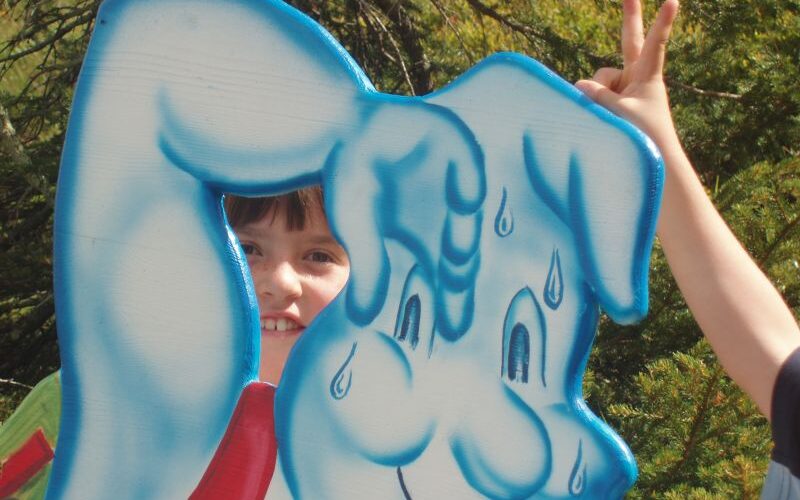 Ein Kind schaut durch den Ausschnitt einer Zeichentrickfigur, die Eiscreme isst, während ein anderes Kind hinter dem Ausschnitt spielerisch die Geste „Hasenohren“ macht.