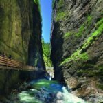 Eine schmale Schlucht mit steilen Felsen, grüner Vegetation und einem türkisfarbenen Fluss; man geht auf einem Holzpfad, der an der Felswand befestigt ist.