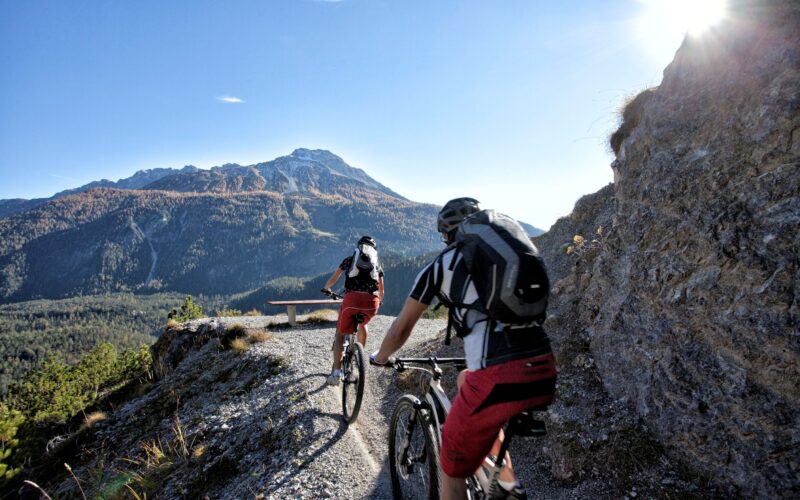 Zwei Radfahrer mit Helmen und Rucksäcken fahren mit Mountainbikes auf einem schmalen Schotterweg mit einem Berg und blauem Himmel im Hintergrund.