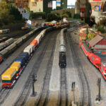 Modelleisenbahnanlage mit mehreren Gleisen, Güterzügen, Gebäuden, Fahrzeugen und Miniaturfiguren, die in einer Halle aufgebaut ist, während die Menschen im Hintergrund zusehen.