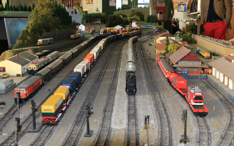 Modelleisenbahnanlage mit mehreren Gleisen, Güterzügen, Gebäuden, Fahrzeugen und Miniaturfiguren, die in einer Halle aufgebaut ist, während die Menschen im Hintergrund zusehen.