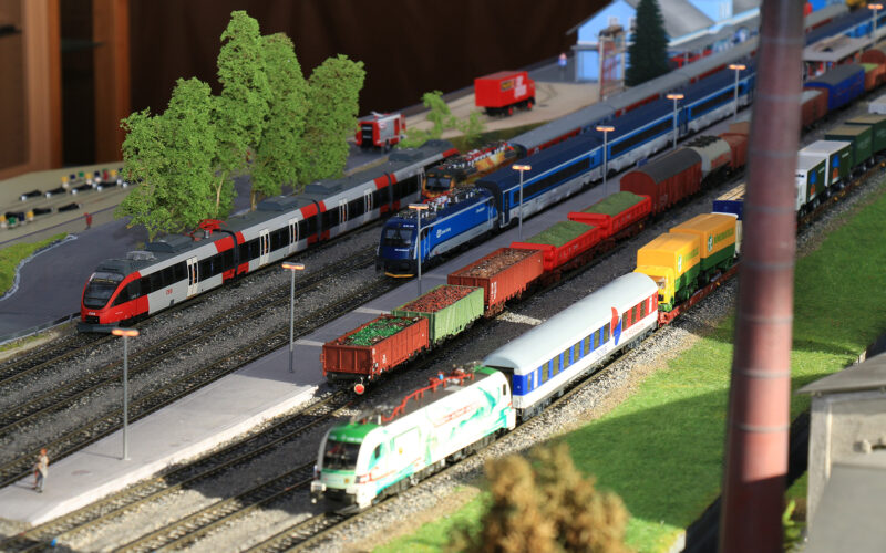 Eine detaillierte Modelleisenbahnanlage mit verschiedenen bunten Lokomotiven und Güterwagen auf mehreren Gleisen, mit Miniaturbäumen und Gebäuden im Hintergrund.