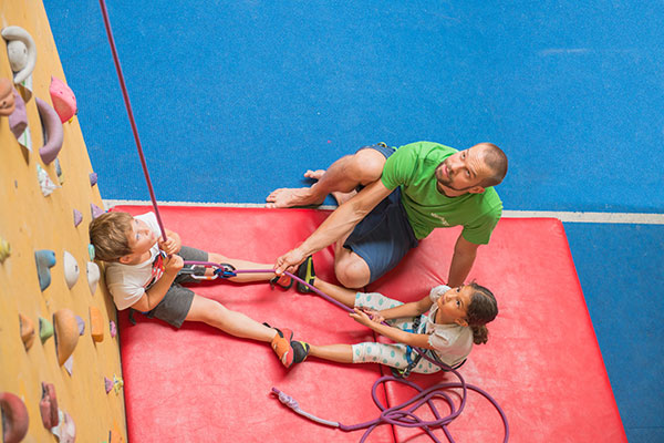 Ein Erwachsener beaufsichtigt zwei Kinder an einer Kletterwand und hilft ihnen mit Seilen, während sie auf einer rot gepolsterten Matte sitzen.