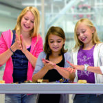 Drei Mädchen stehen lächelnd an einem Tisch und formen in einem hell erleuchteten Raum mit ihren Händen Ton oder Teig.