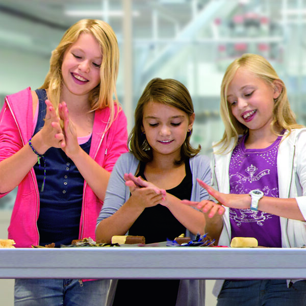 Drei Mädchen stehen lächelnd an einem Tisch und formen in einem hell erleuchteten Raum mit ihren Händen Ton oder Teig.