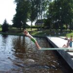 Eine Person in grüner Badehose springt von einem Sprungbrett in einen See, während andere zusehen und sich auf einem hölzernen Steg und einer Grasfläche in der Nähe entspannen.