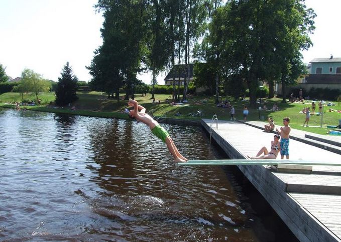 Eine Person in grüner Badehose springt von einem Sprungbrett in einen See, während andere zusehen und sich auf einem hölzernen Steg und einer Grasfläche in der Nähe entspannen.
