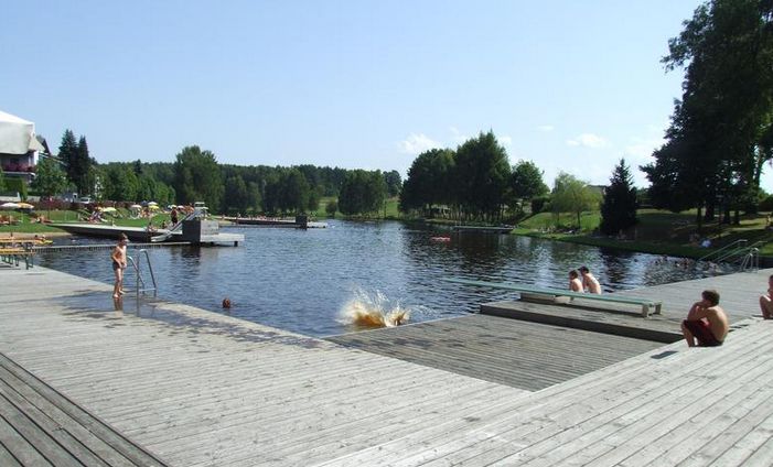 Auf einer von Bäumen umgebenen Holzterrasse am Seeufer kann man sich entspannen und schwimmen, während eine Person ins Wasser springt und es dabei ordentlich krachen lässt.