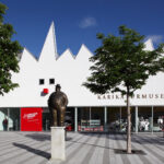 Weißes, modernes Gebäude mit gezacktem Dach und der Aufschrift "Karikaturmuseum"; Statuen auf Sockeln und Bäume säumen den gestreiften Platz davor unter einem klaren blauen Himmel.