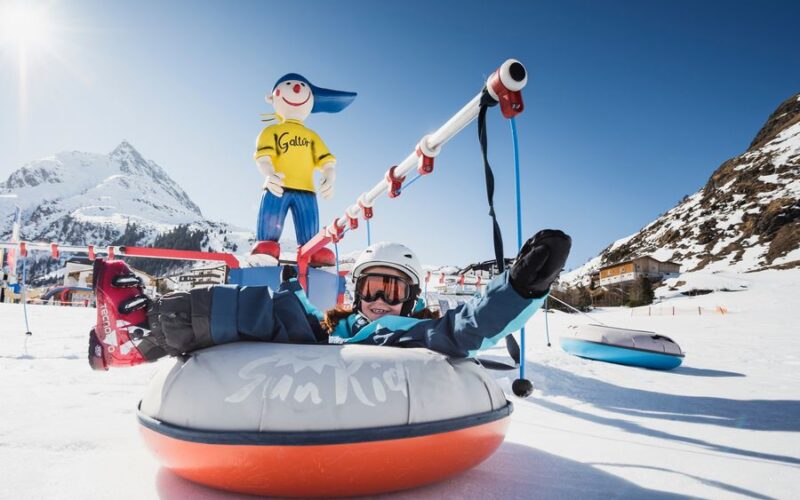 Eine Person in Winterkleidung fährt auf einem aufblasbaren Schneeschlauch und hält sich dabei an einem Zugseil fest. Im Hintergrund sind ein verschneiter Berg und eine Zeichentrickfigur zu sehen.