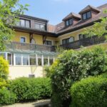 Ein großes gelbes Haus mit einem verglasten Wintergarten, dunklen Holzbalkonen und Dachgauben, umgeben von grünen Sträuchern und Bäumen.