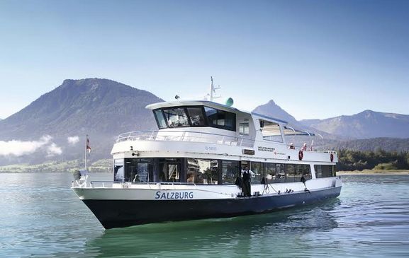 Ein weißes Fahrgastschiff mit dem Namen "Salzburg" fährt auf einem ruhigen See mit Bergen und klarem blauen Himmel im Hintergrund.
