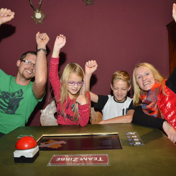 Vier Personen sitzen an einem Tisch mit einem "Team ZIRBE"-Schild und heben feierlich ihre Arme. Ein roter Buzzer, ein Spielbrett und Karten liegen auf dem Tisch.