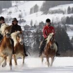 Drei Personen reiten auf Pferden durch Schnee in einer ländlichen, bergigen Gegend mit Häusern und Bäumen im Hintergrund.