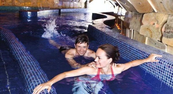 Ein Mann und eine Frau schwimmen in einem Hallenbad mit gefliestem Rand und Steinwänden, wobei der Mann hinter der Frau steht und sich am Beckenrand festhält.