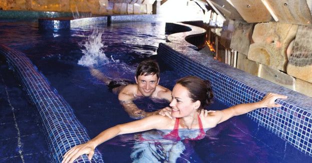 Ein Mann und eine Frau schwimmen in einem Hallenbad mit gefliestem Rand und Steinwänden, wobei der Mann hinter der Frau steht und sich am Beckenrand festhält.