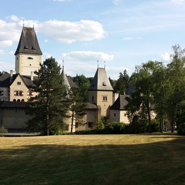 Eine historische Steinburg mit hohen Türmen und steilen Dächern steht umgeben von Bäumen und einer großen Wiese unter einem teilweise bewölkten Himmel.