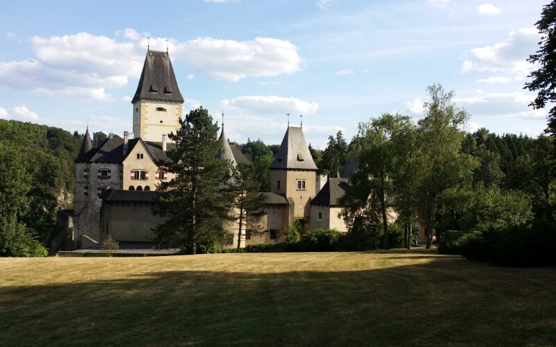 Eine historische Steinburg mit hohen Türmen und steilen Dächern steht umgeben von Bäumen und einer großen Wiese unter einem teilweise bewölkten Himmel.