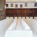 Ein langer Esstisch mit weißen Stühlen und Tischtüchern, der für eine formelle Veranstaltung gedeckt ist, mit Holzvertäfelung und dekorativen Wappen an der Wand im Hintergrund.