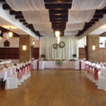 Ein Bankettsaal mit geschmückten Tischen und Stühlen, weißen Deckenvorhängen, Holzböden, Backsteinsäulen und einem mit Grünzeug und Kränzen geschmückten Haupttisch am Ende.