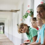 Vier Kinder sitzen auf einer Bank in einem hellen Korridor mit weißen Wänden und rosa Blumen und schauen in die Kamera.