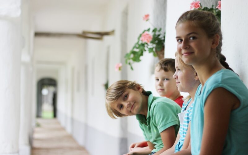 Vier Kinder sitzen auf einer Bank in einem hellen Korridor mit weißen Wänden und rosa Blumen und schauen in die Kamera.
