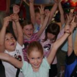 Eine Gruppe von Kindern steht dicht beieinander in einem Haus, hebt die Arme und lächelt in die Kamera. Im Hintergrund sind Bowlingkugeln zu sehen.