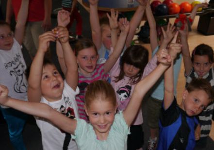 Eine Gruppe von Kindern steht dicht beieinander in einem Haus, hebt die Arme und lächelt in die Kamera. Im Hintergrund sind Bowlingkugeln zu sehen.